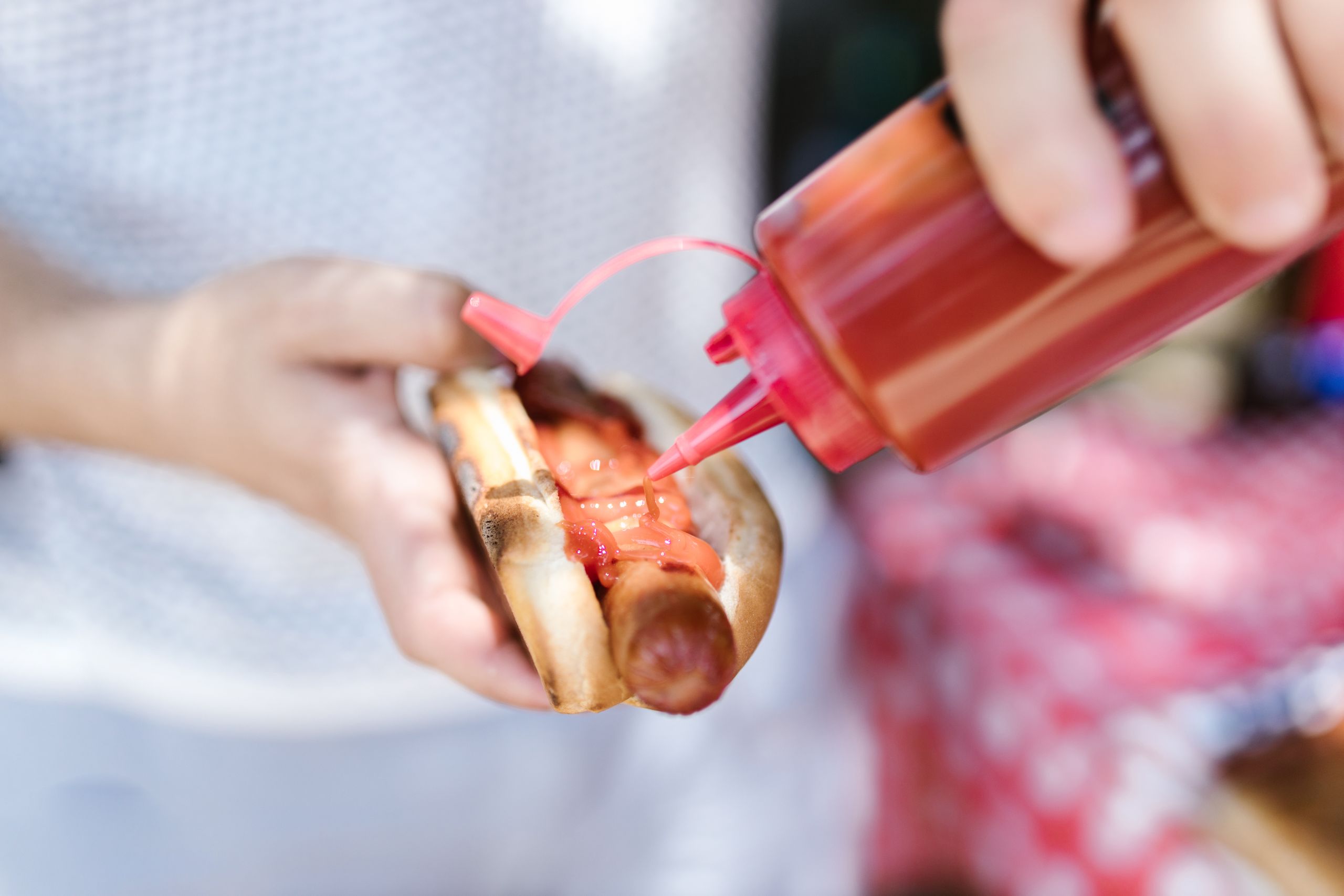 Person adding ketchup to a grilled hot dog held in hands outdoors.